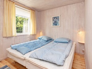 A single bed with a quilted blue cover, a window with yellow curtains, and a framed picture on the wall.