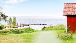 Un sentier bordé d’herbe mène à un bâtiment rouge, situé au bord d’une étendue d’eau calme, avec une jetée qui s’avance sur l’eau.