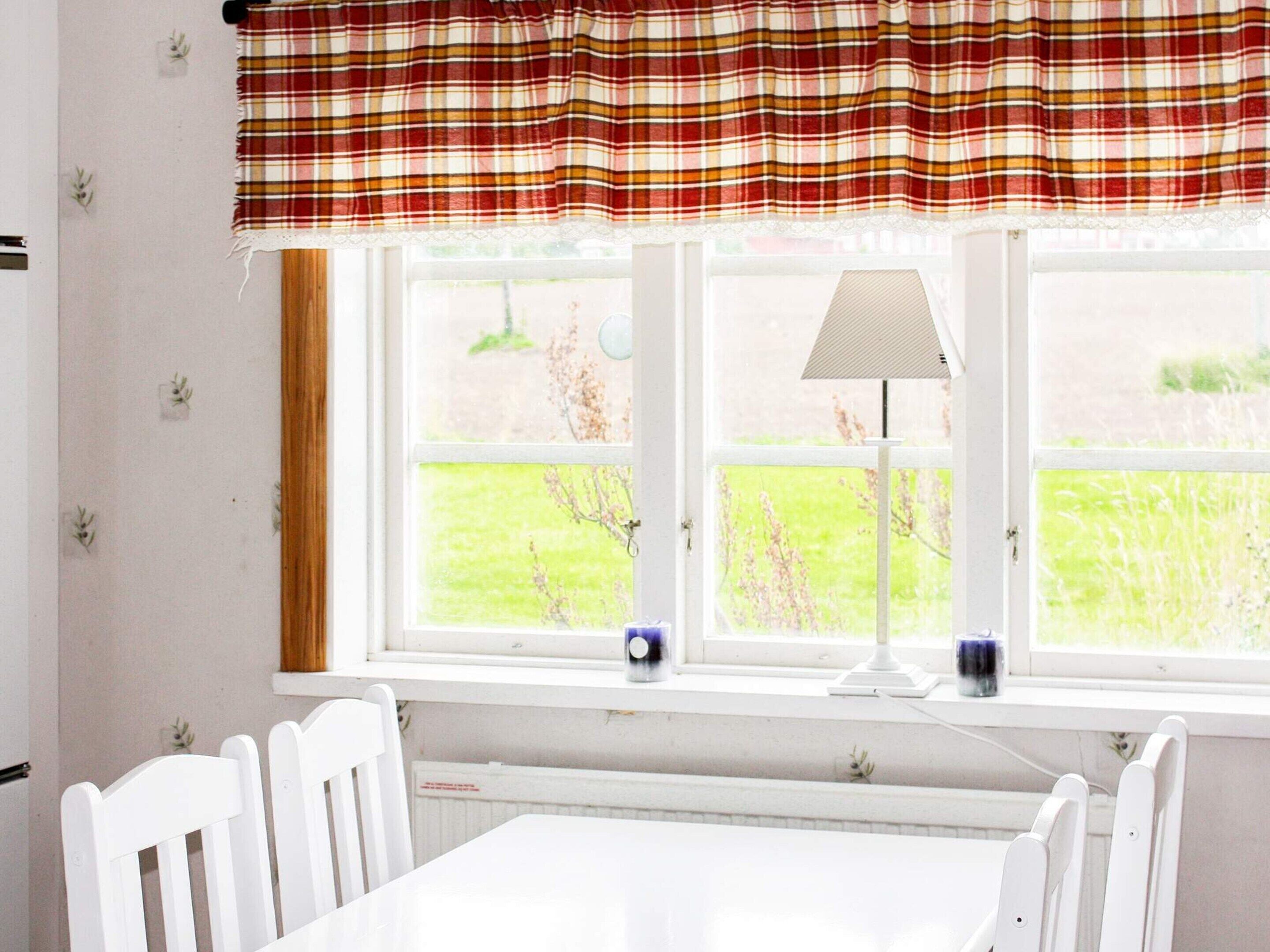 A room with a white table and chairs, a window with a view of greenery, and a lamp on the windowsill.