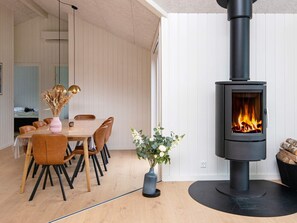 Ein modernes Interieur mit einem zentral platzierten Esstisch, Stühlen, einer Vase mit Trockenpflanzen und einer blauen Vase mit Blumen sowie einem Kaminofen.