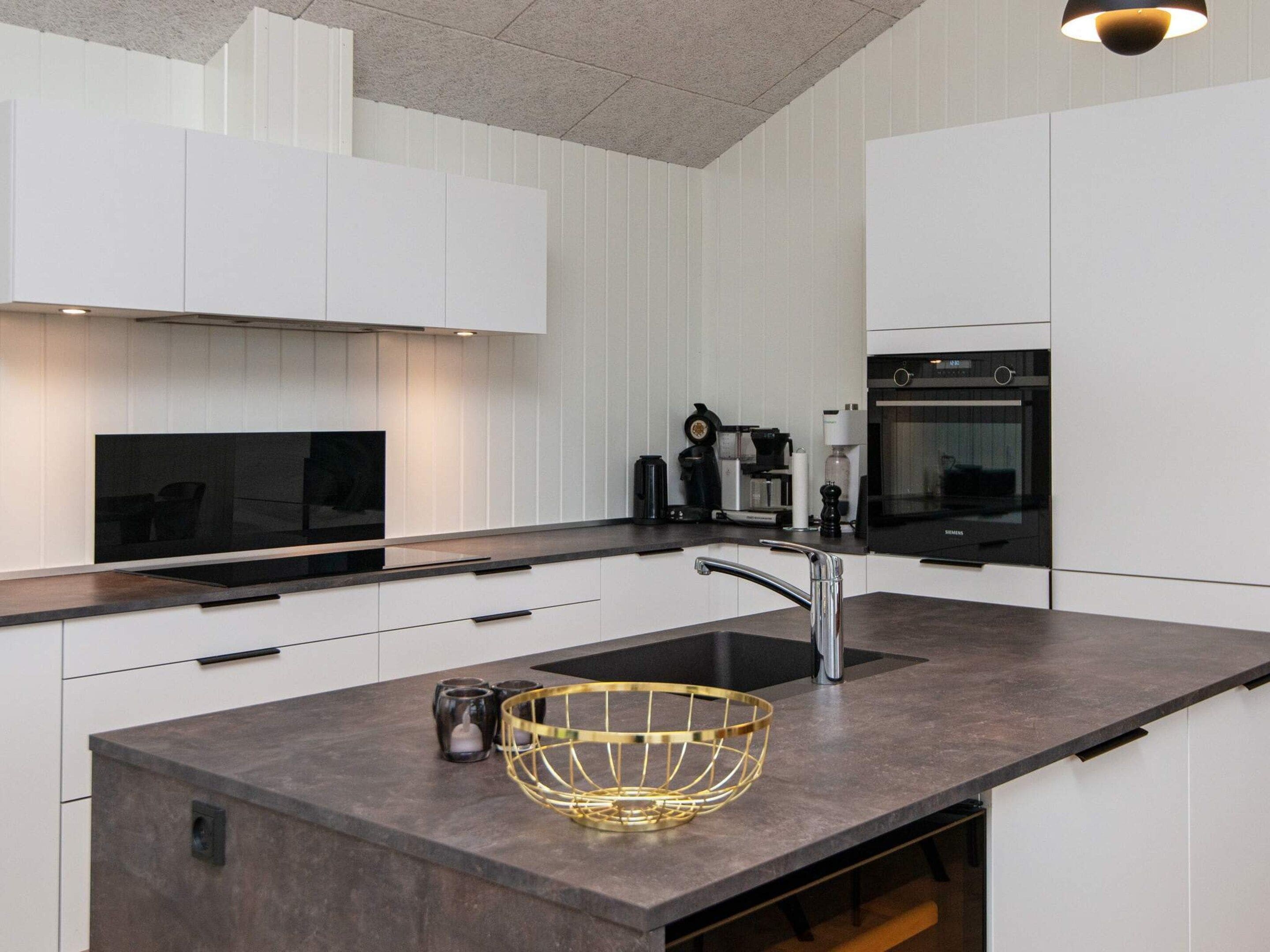 Una cocina moderna con armarios blancos, encimera oscura, horno integrado y una isla grande con cesto de alambre dorado.
