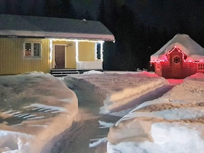 A snow-covered landscape with two buildings, one with a porch and another with red lights.