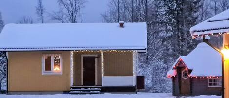 Uma casa coberta de neve com varanda e um pequeno galpão iluminado.