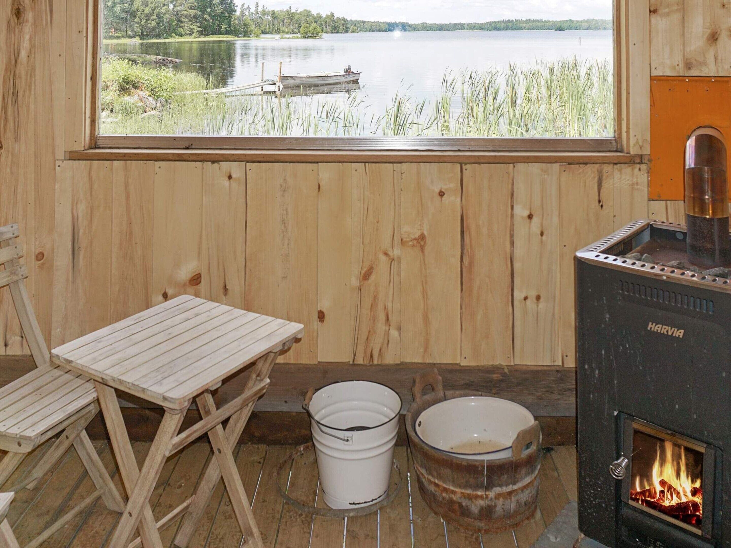 A wooden cabin with a view of a lake, a Harvia wood stove, a table, two buckets, and a chair.