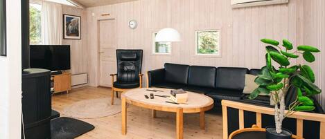 Ein modernes Wohnzimmer mit Holzboden, einem schwarzen Ledersofa, einem Holztisch, einem schwarzen Ledersessel, einem Fernseher und einer Zimmerpflanze.