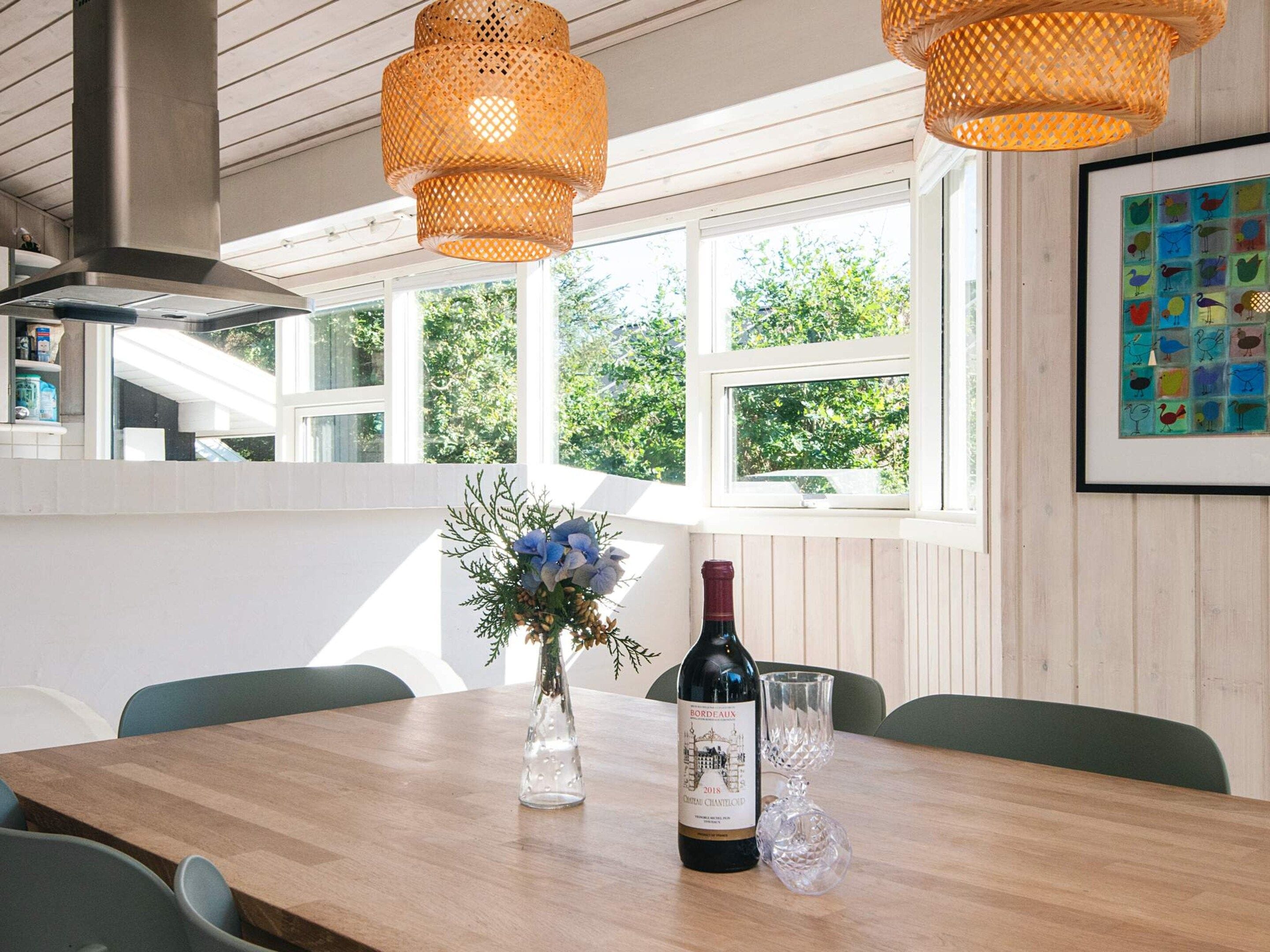 A dining area with a wooden table, two wicker pendant lights, a bottle of wine, and a vase of flowers.