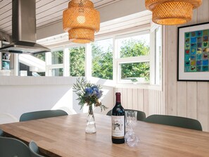 A dining table with a bottle of wine, two glasses, and a floral arrangement in a room with wooden walls, large windows, and woven light fixtures. - 4 Star Holiday Home in Blavand (Blavand)