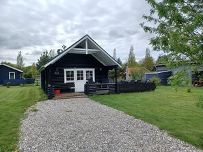 A small, single-story cabin with a white door and windows, surrounded by a gravel driveway and grassy areas. - 6 Person Holiday Home in Hovborg (Hovborg)