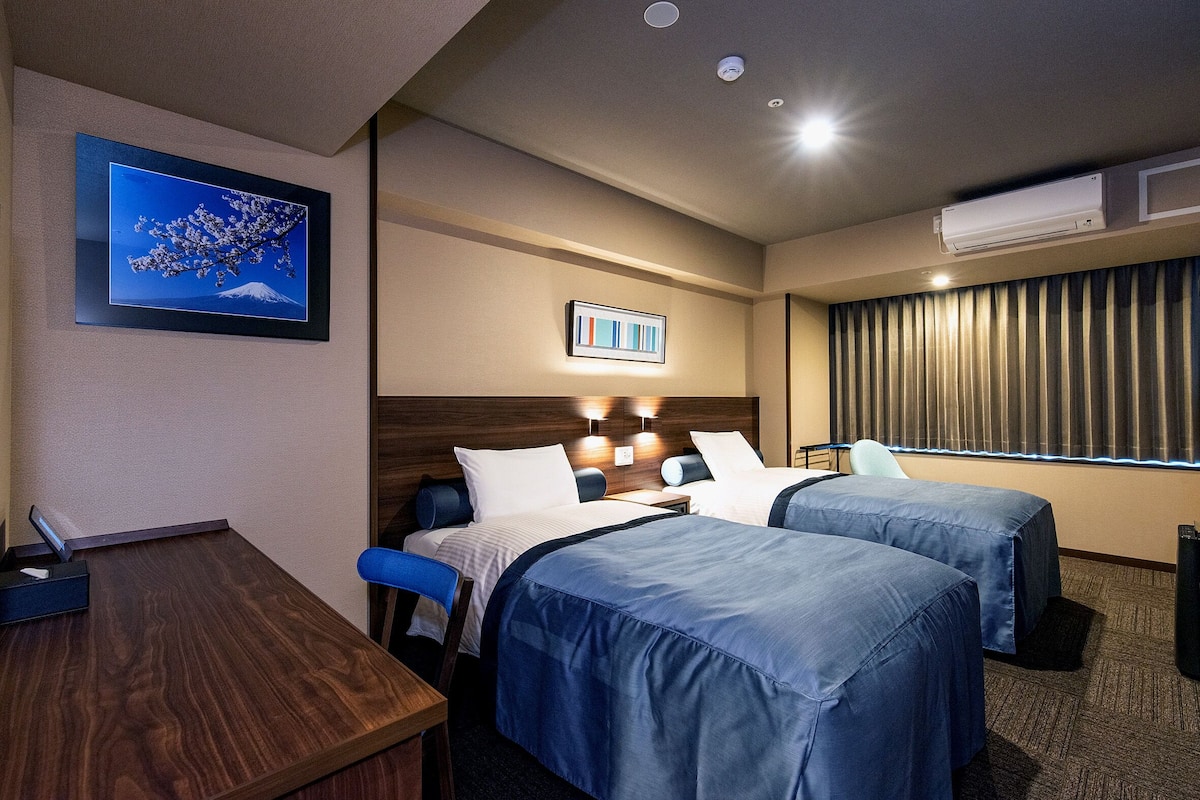 A hotel room with two beds, a desk, a chair, a framed picture, and a window with curtains.