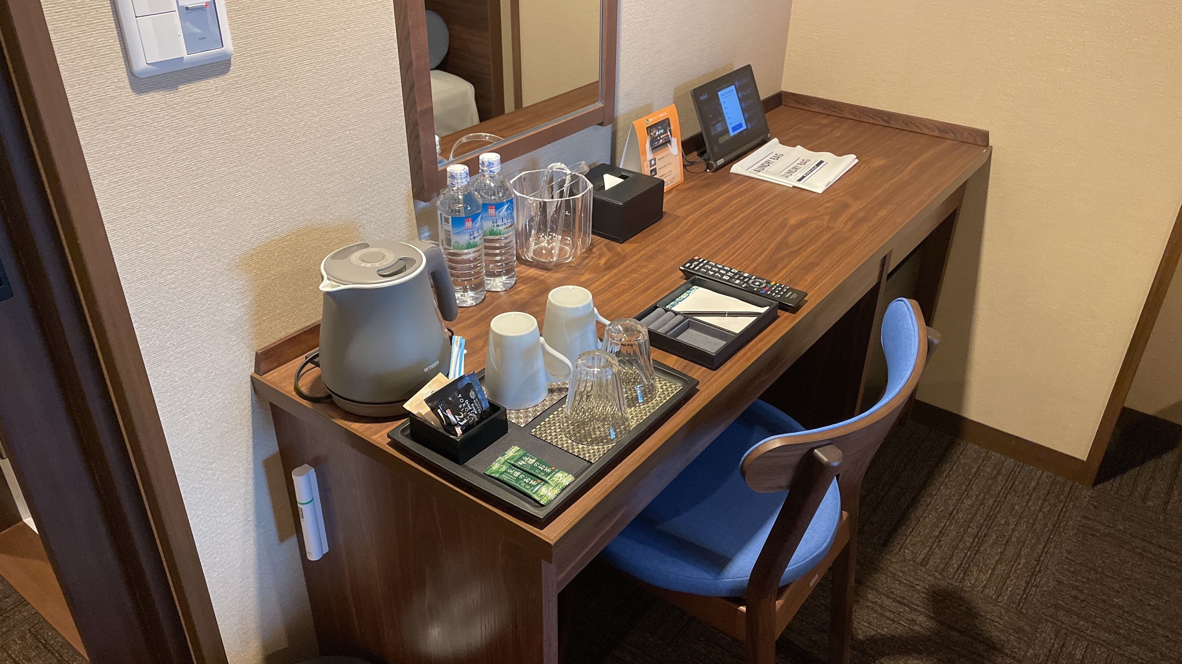 Escritorio de hotel con tetera, botellas de agua, vasos, control remoto y una bandeja con aperitivos.