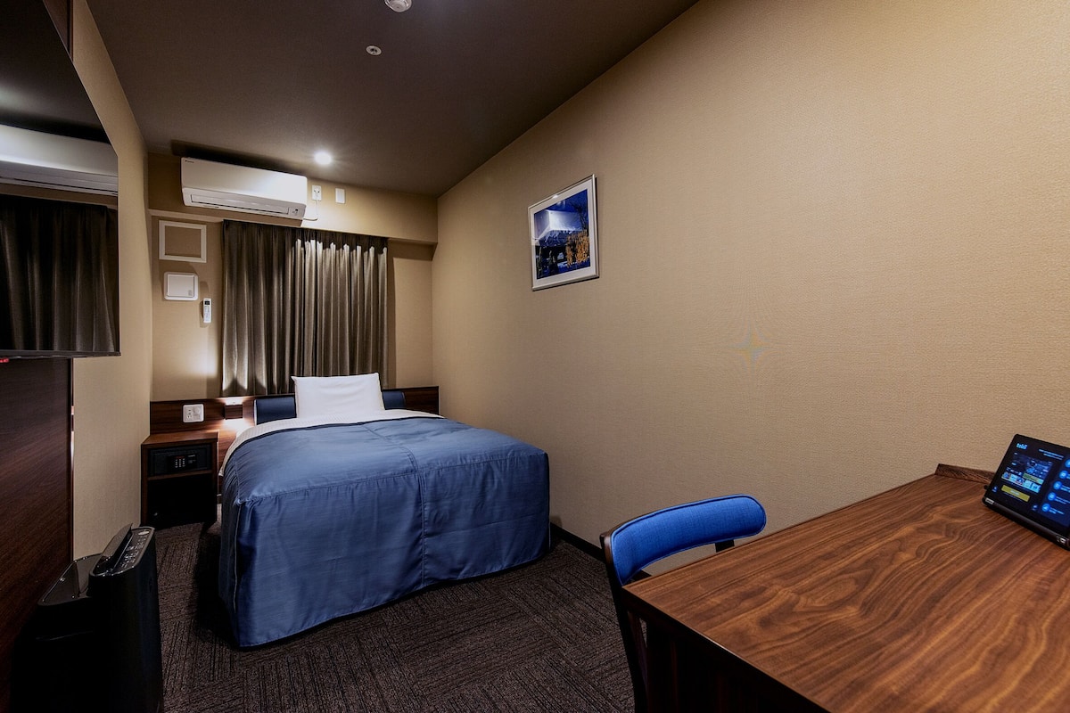 A hotel room with a bed, a desk, a chair, an air conditioner, a picture on the wall, and a control panel on the desk.