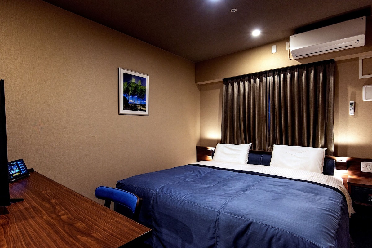 A hotel room with a neatly made bed, a desk with a computer, a framed picture on the wall, and an air conditioning unit.
