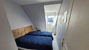 A bedroom with a sloped ceiling, a wooden headboard, two beds with blue bedding, a radiator, and a window with a view of the outside.