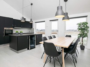 Une cuisine moderne avec des meubles de couleur noire, une table à manger en bois et des chaises noires. Des luminaires suspendus sont placés au-dessus de la table, et une plante en pot se trouve sur le rebord de la fenêtre.
