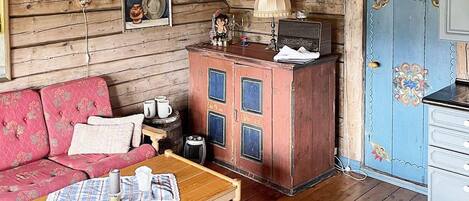 A cozy cabin interior with a wooden ceiling, a red cabinet with blue accents, a pink sofa, and a small table with a plaid tablecloth.