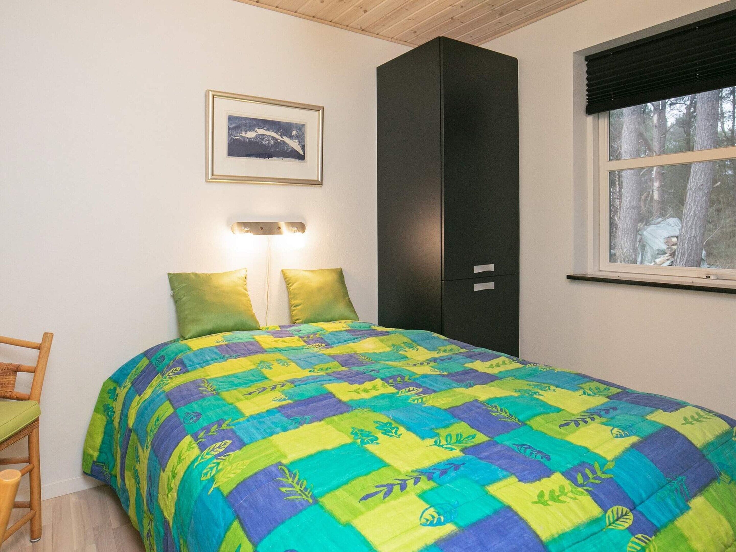 A bedroom with a bed, two bedside lamps, a framed picture on the wall, a wardrobe, and a window with a view of trees.