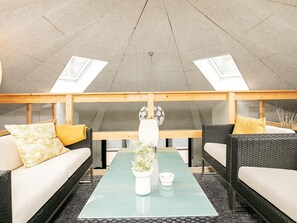 A modern, open-plan living space with high ceilings and skylights, featuring a central glass coffee table, wicker furniture, and a minimalist decor. - 4 Star Holiday Home in Laeso (Læsø)