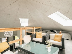 A modern, open-plan living space with a high ceiling and skylights, featuring a glass coffee table, wicker furniture, and a small white vase with dried flowers.