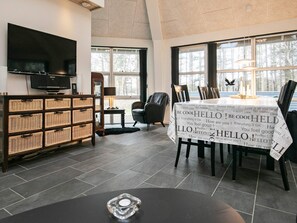 A modern dining area with a table covered in a 'Hello' themed tablecloth, a television, wicker baskets, and a view of trees through large windows. - 4 Star Holiday Home in Laeso (Læsø)