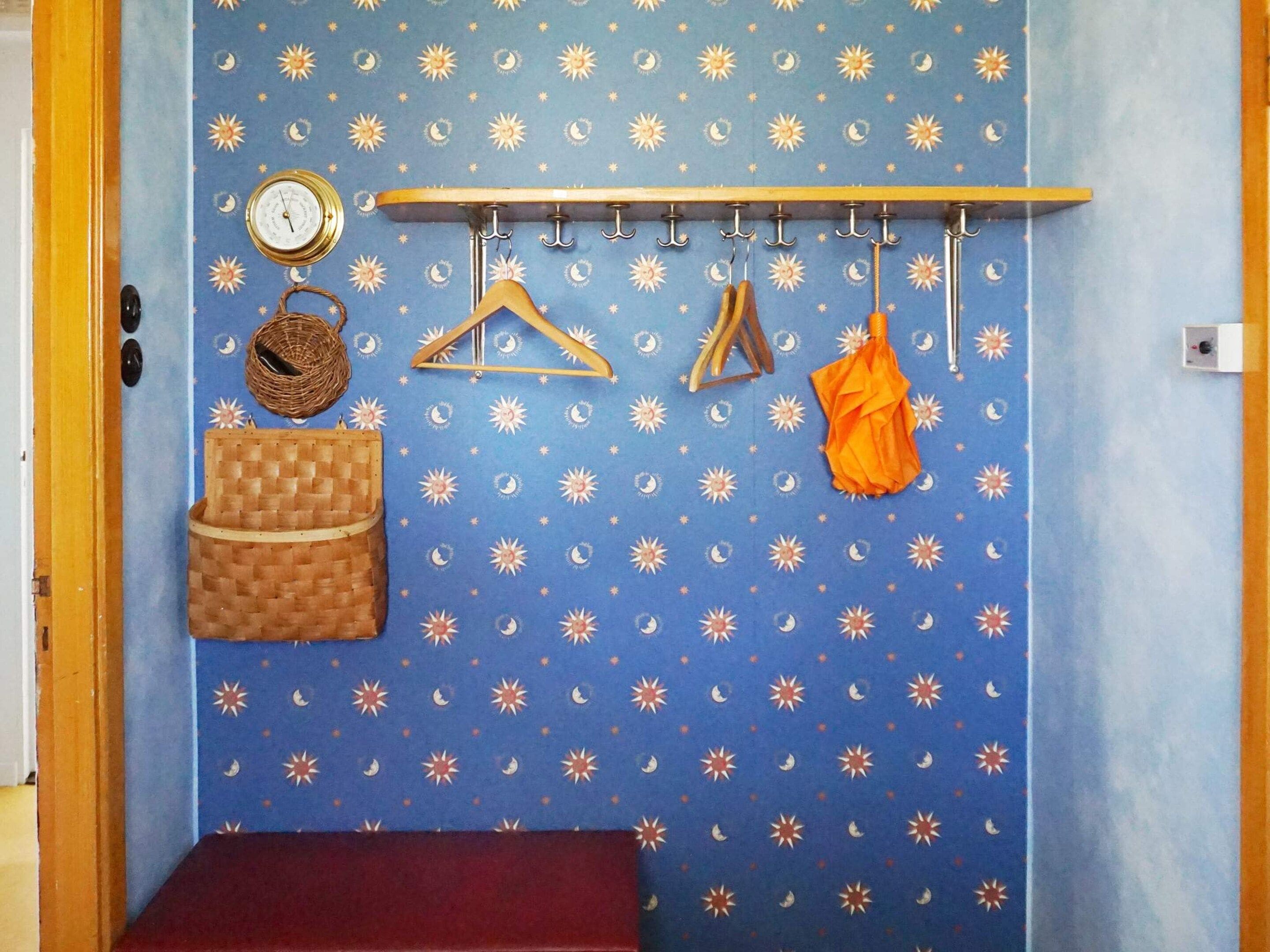 Une pièce avec un mur bleu orné d’un motif représentant une étoile et une lune, un porte-manteau en bois avec des cintres, une petite horloge ronde, un panier en osier et un banc.