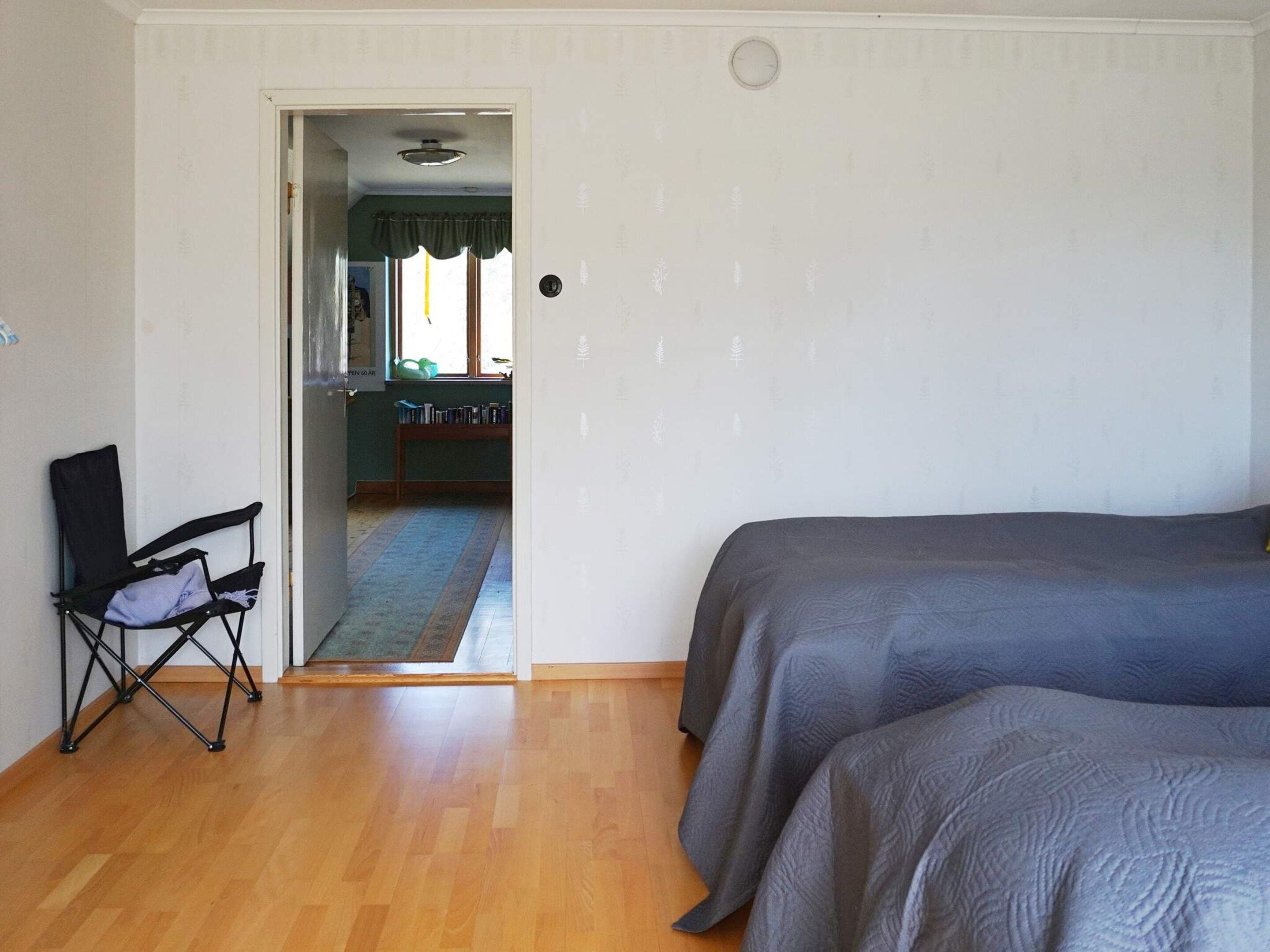 Une pièce avec un plancher en bois, une chaise pliante noire et deux lits avec du linge de lit gris.
