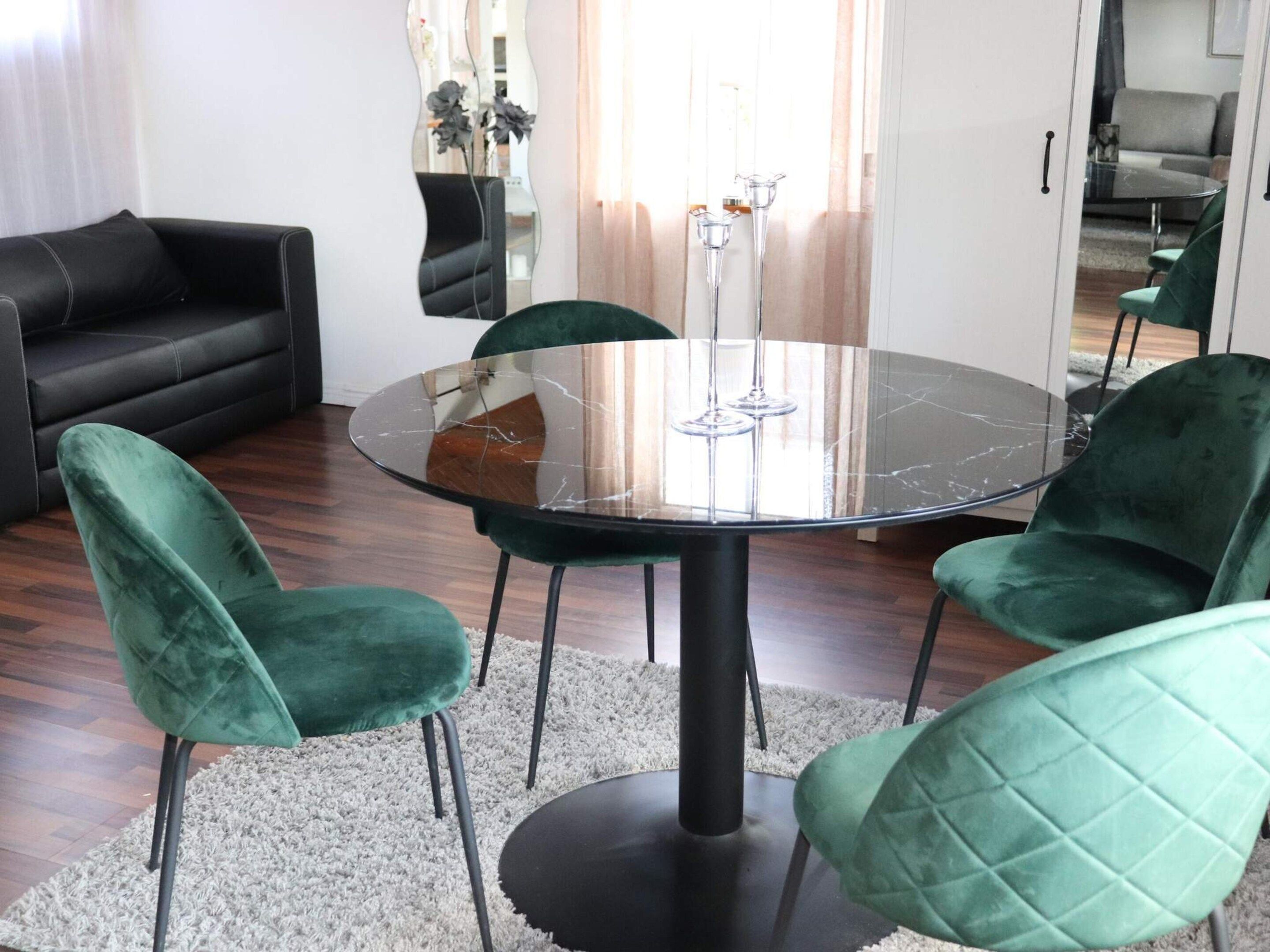 A round glass table with four chairs, a black sofa, and a white rug in a room with wooden flooring.