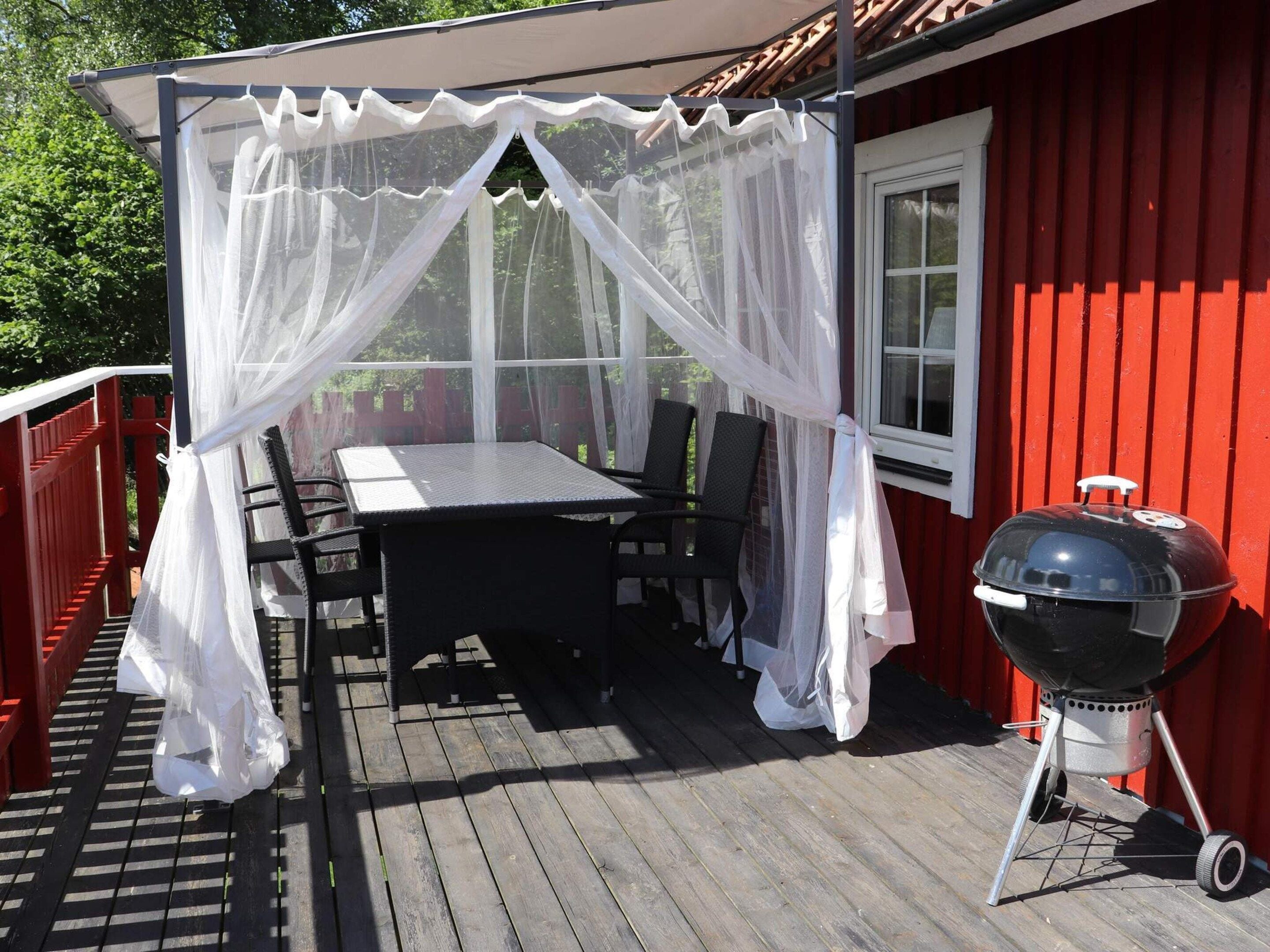 A covered outdoor dining area with a table and chairs, a barbecue grill, and a red building in the background.