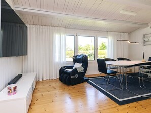 A modern room with a wooden floor, a black massage chair, a dining table with chairs, and large windows with curtains. - 4 Person Holiday Home in Faxe Ladeplads (Faxe Ladeplads)