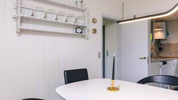 Ein Essbereich mit einem weißen Tisch, schwarzen Stühlen, einem Kerzenhalter und einem an der Wand befestigten Regal mit Tellern.