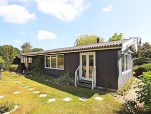 A small, dark wooden house with a porch, large windows, and a garden with stepping stones.
