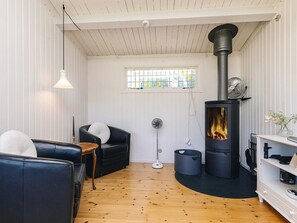 A cozy living room with a fireplace, a black leather sofa, a white armchair, and a wooden floor. - 4 Person Holiday Home in Faxe Ladeplads (Faxe Ladeplads)