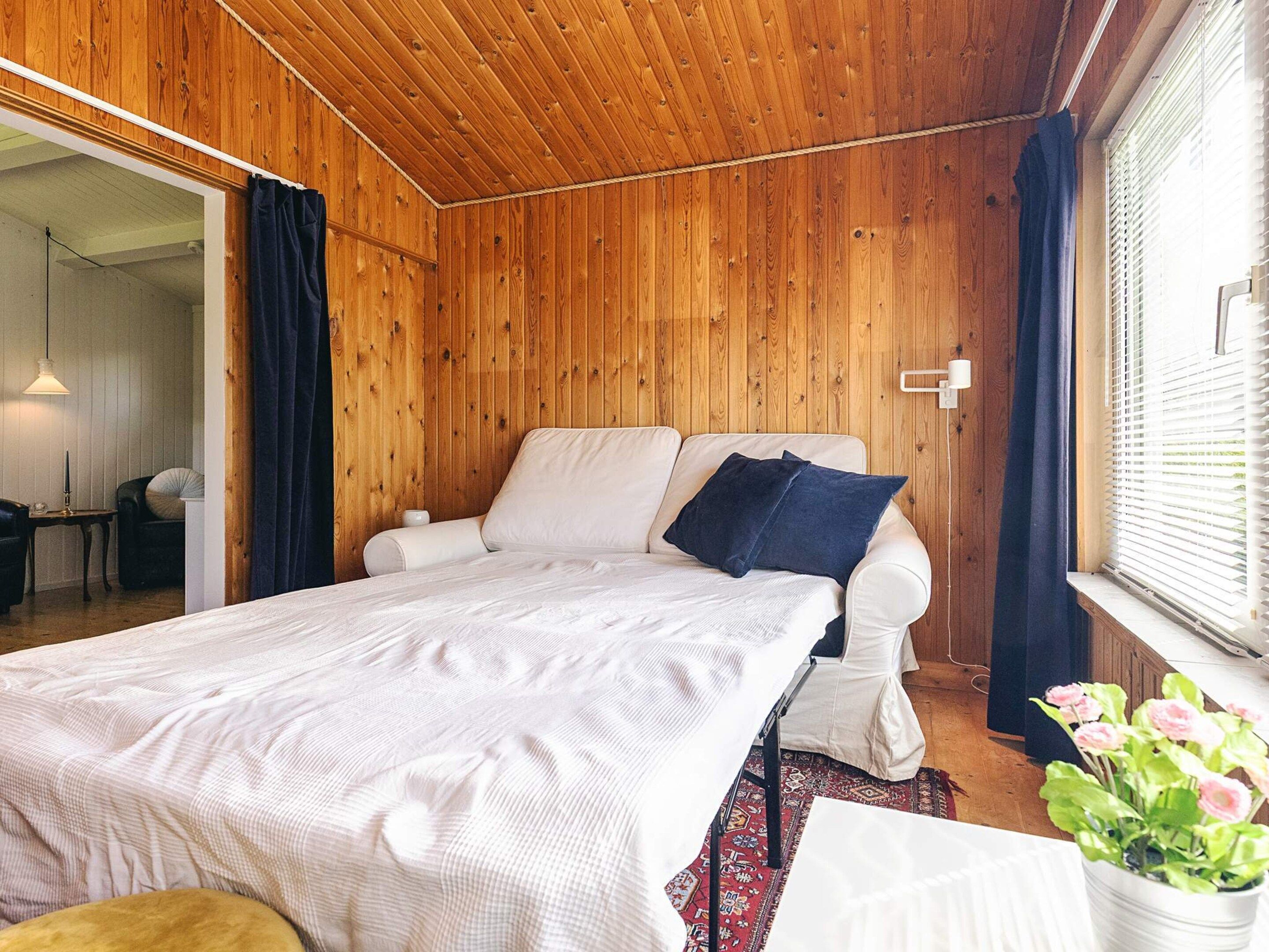 A bedroom with a wooden ceiling, a bed with white bedding, a dark blue pillow, a small table with a lamp, and a window with blinds.