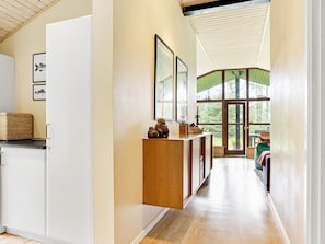 A narrow hallway with a wooden cabinet, framed artwork, and a large window leading to an outdoor area.