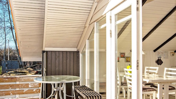 Een houten terras met een glazen tafel en stoelen, een bankje, en een schuifpui die leidt naar een eethoek binnen met witte meubels.
