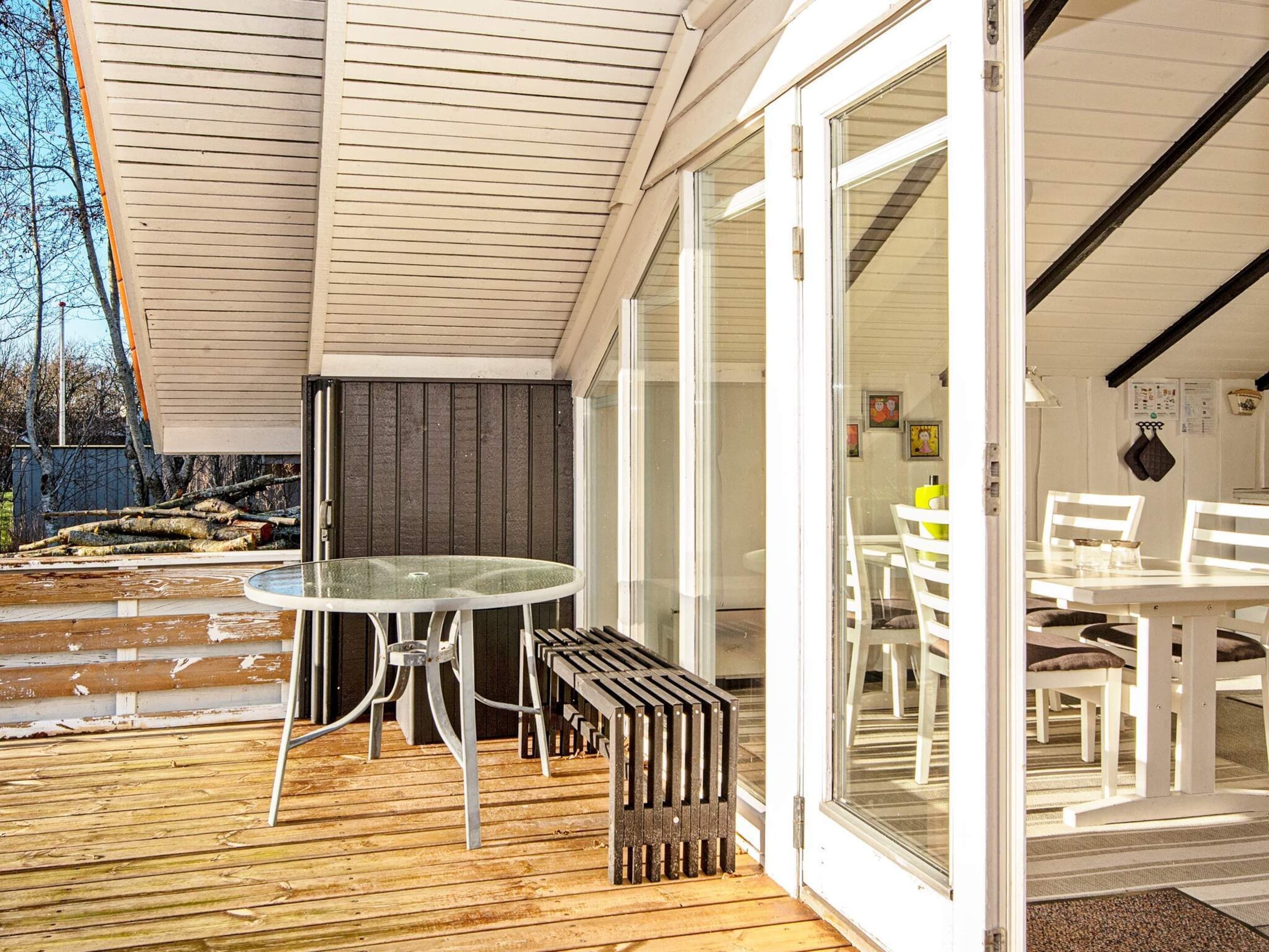 Una terraza abierta con una mesa y sillas de vidrio, piso de madera y vistas al interior a través de puertas de cristal.