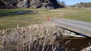 Um campo de golfe com um afloramento rochoso ao fundo, uma ponte sobre a água e uma pessoa caminhando à distância.