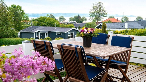 Een houten terras met een tafel, stoelen, een bloempot en uitzicht op huizen en bomen.