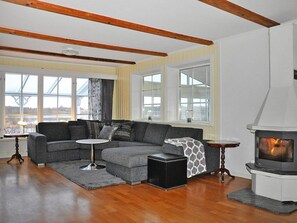 A modern living room with a sectional sofa, a small round table, a black ottoman, a fireplace, and large windows.