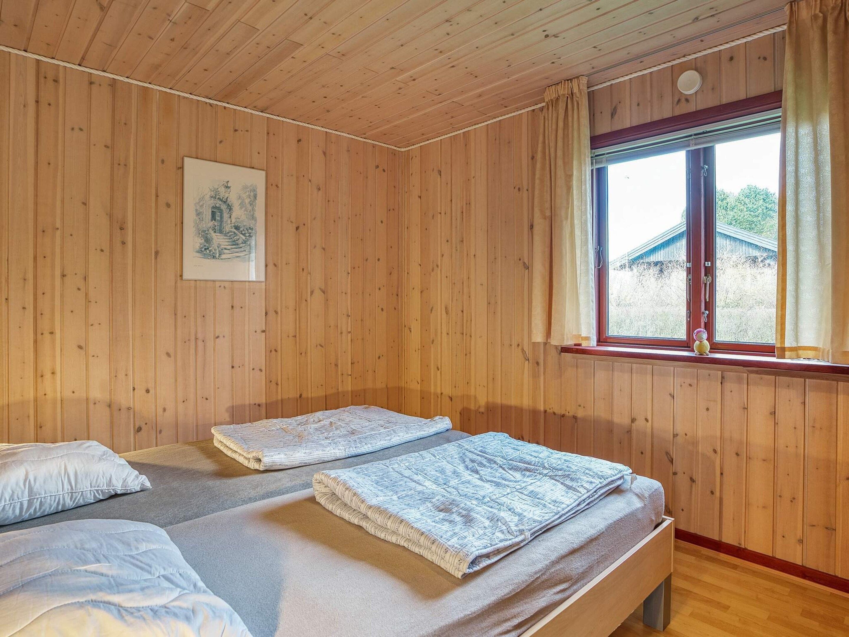 A room with wooden walls, a window with curtains, two beds with pillows, and a framed picture on the wall.