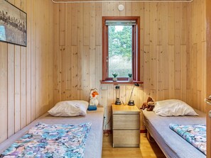 Una habitación pequeña y acogedora con paredes de madera, dos camas individuales con ropa de cama estampada, una mesita de noche con lámparas y una ventana con vistas a la vegetación.