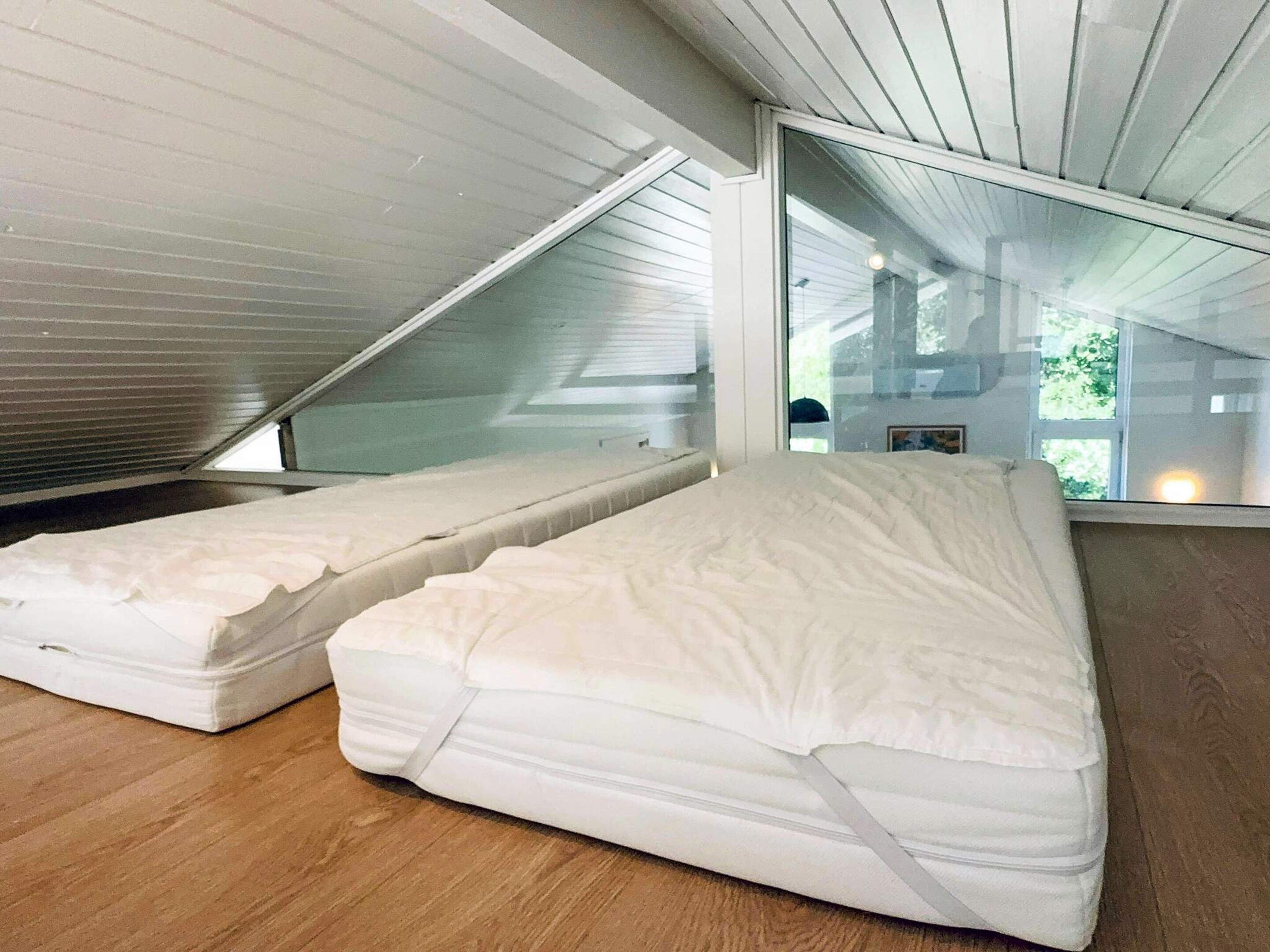 A room with two mattresses on the floor, a sloped ceiling, and a large window.