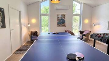 A room with a ping pong table, foosball table, and a large window with a view of greenery.