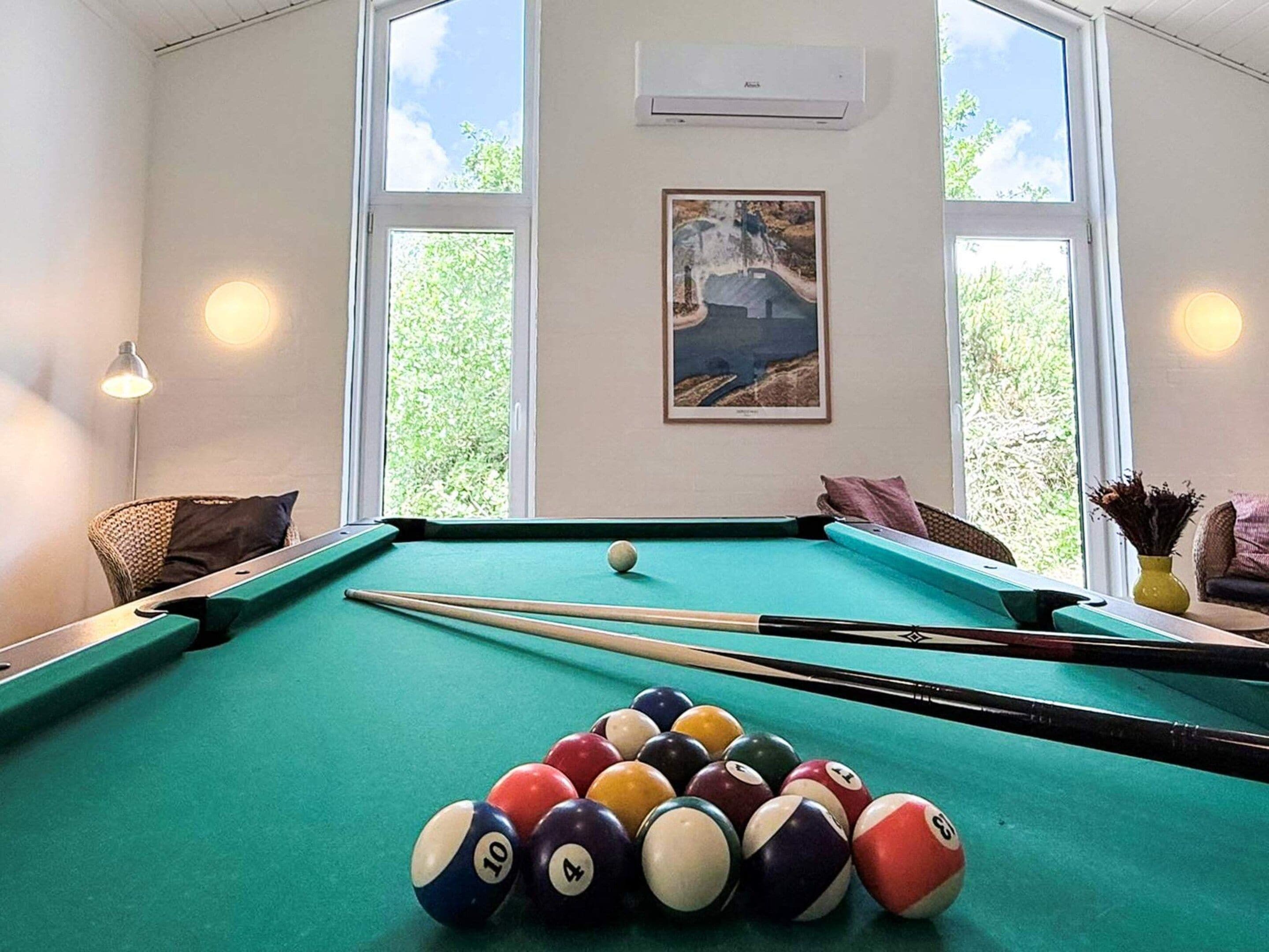 A pool table with balls, a framed picture, and a window with a view of greenery.