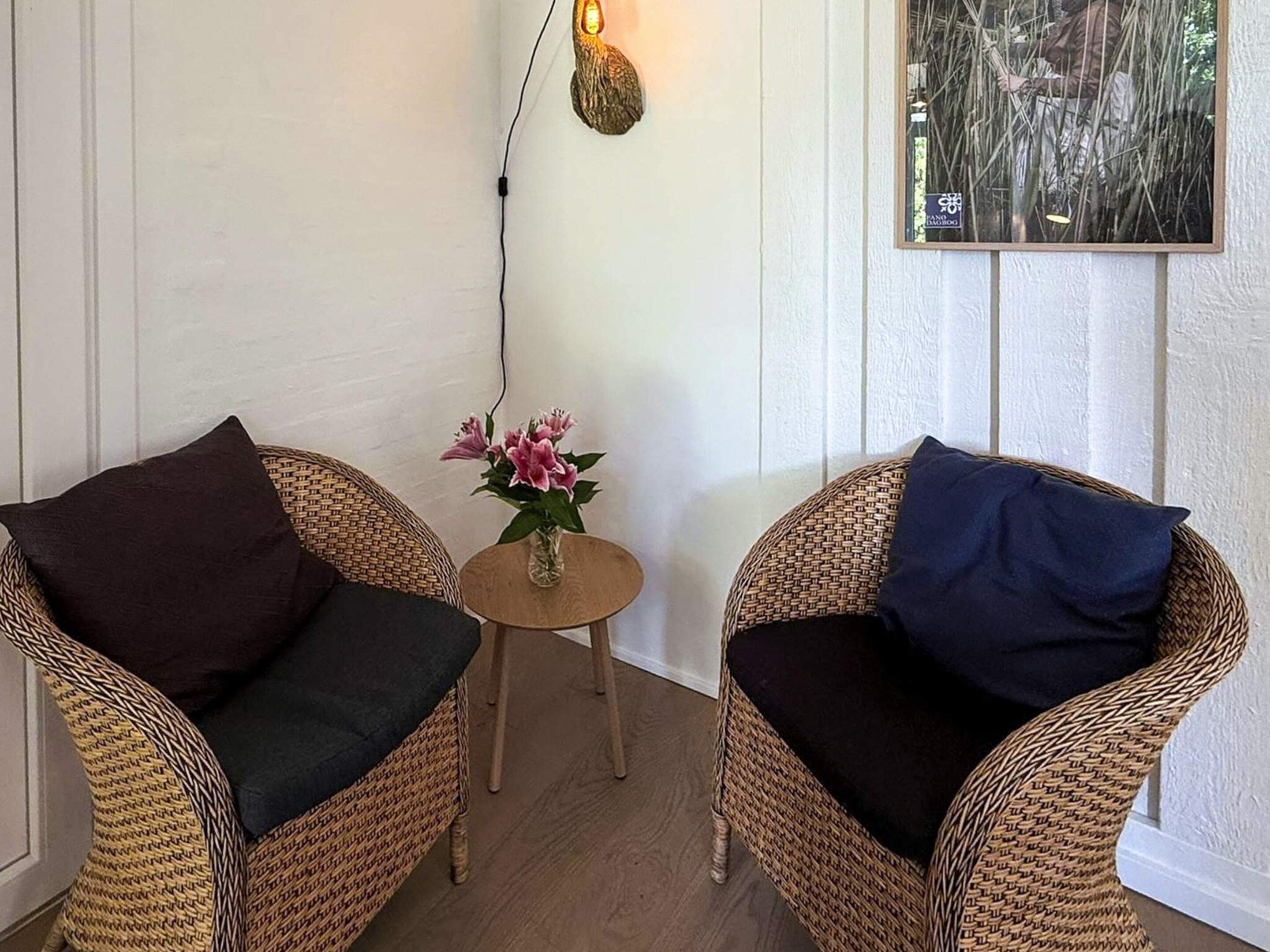 Two wicker chairs with dark cushions, a small wooden table with a vase of flowers, and a wall-mounted light fixture.