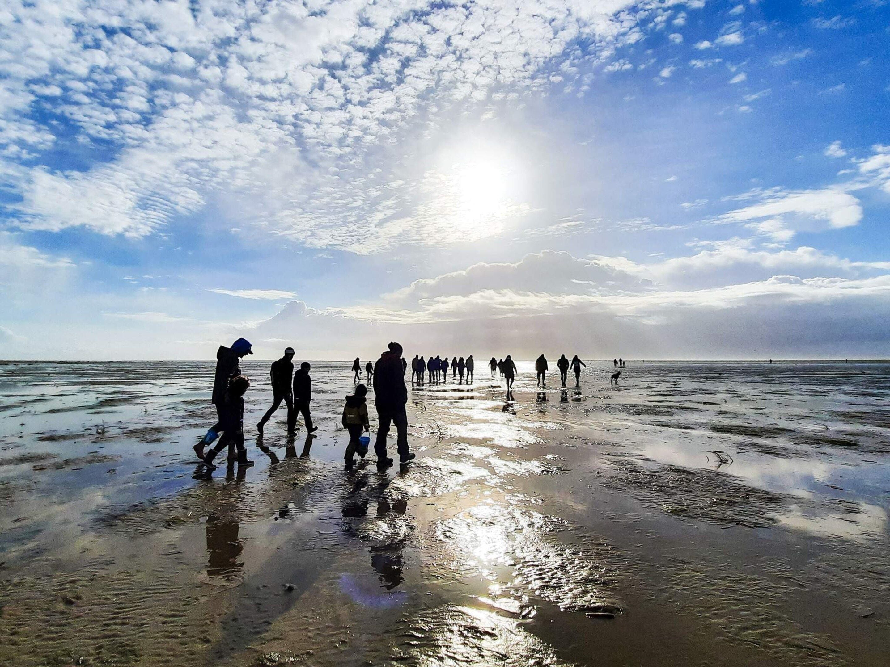 Una spiaggia con persone che camminano sulla sabbia bagnata, il sole visibile nel cielo e il mare sullo sfondo.