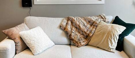 Sala de estar con un sofá, almohadas, una manta, una mesa de centro blanca y un ventanal con vistas a zonas verdes.