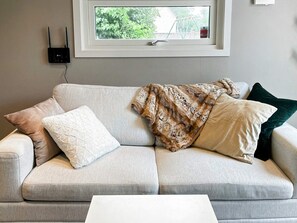 A living room with a sofa, pillows, a blanket, a white coffee table, and a window with a view of greenery.