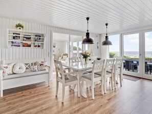 A dining area with a white table and chairs, a white bench with floral cushions, and a view of the sea through large windows.