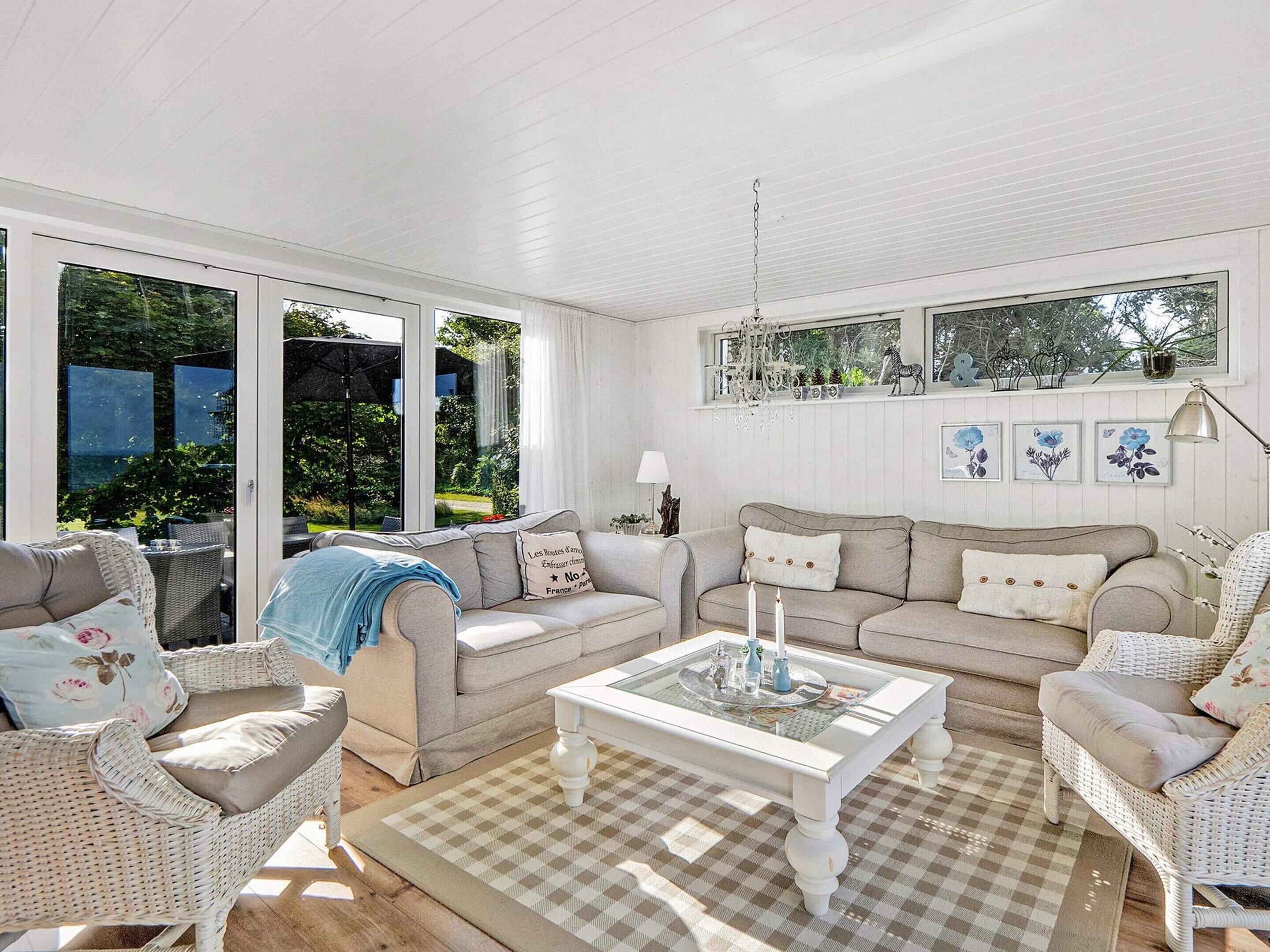 A sunlit living room with a checkered rug, two sofas, a coffee table, wicker chairs, and a view of greenery through large windows.