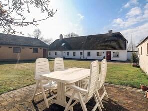 A patio with a table and chairs, a grassy lawn, and a white building with a red door in the background. - 4 Star Holiday Home in Aabybro (Aabybro)